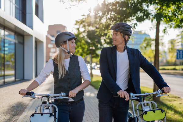 Colleagues commuting through the city, talking and walking by bike on street. Middle-aged city commuters traveling from work by bike after a long workday. Husband and wife riding bikes in city.