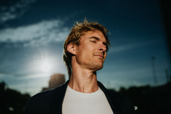 Portrait of handsome mature man with blond hair and closed eyes, standing outdoors in the city, in front of reflective wall.