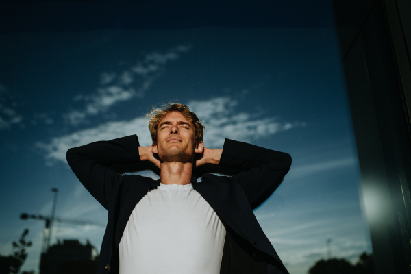 Portrait of handsome mature man with blond hair and closed eyes, standing outdoors in the city, in front of reflective wall.