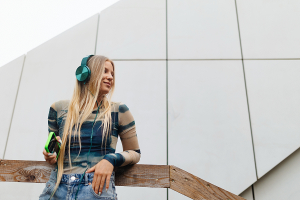 Portrait of a Generation Z girl student listening to music via headphones outdoors in the city. Student spending free time online and alone. Concept of gen Z as loneliest generation.