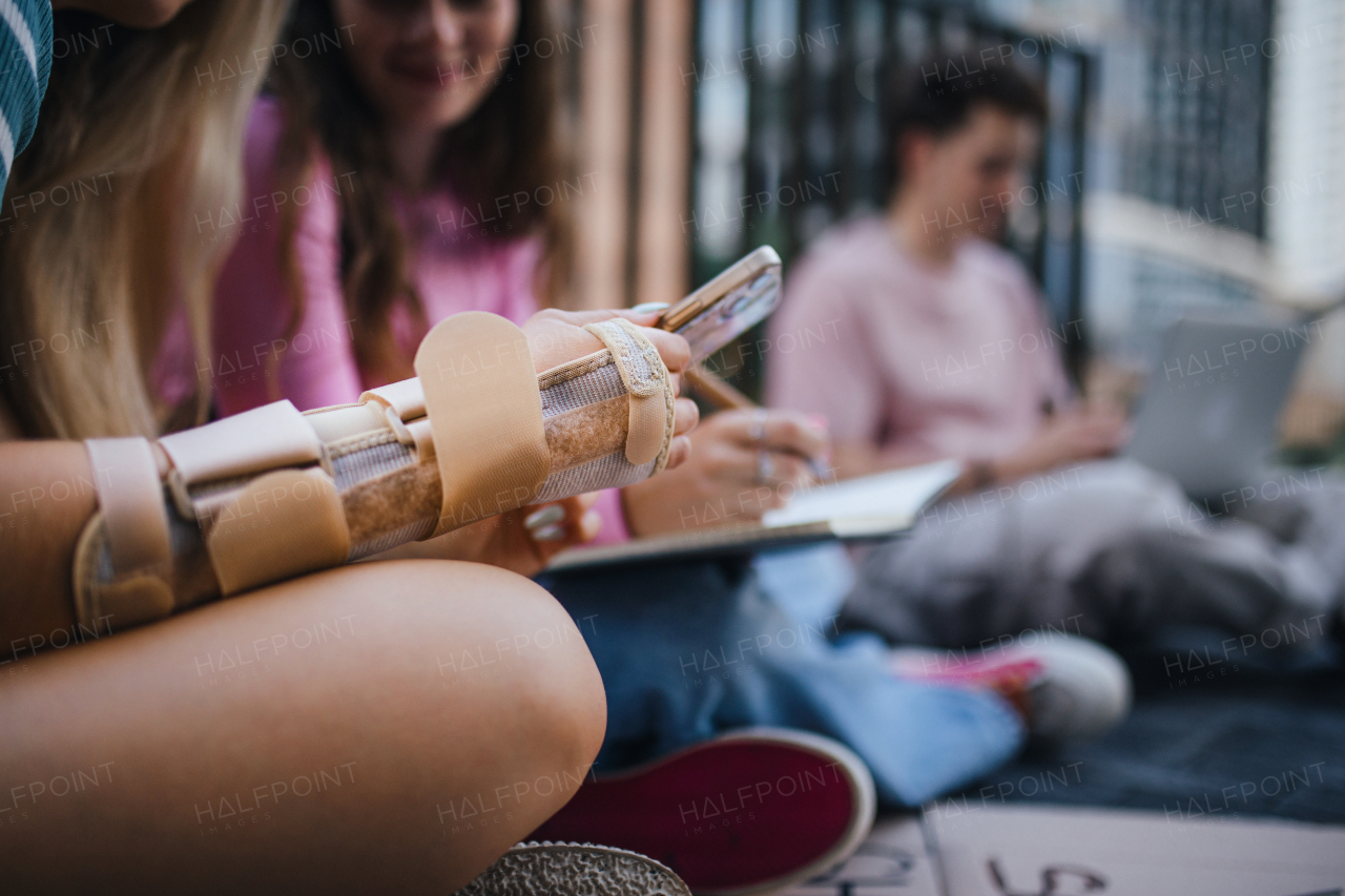 Close up of broken hand of gen Z student studying with firnends outdoors after school. Arm in support forearm brace.