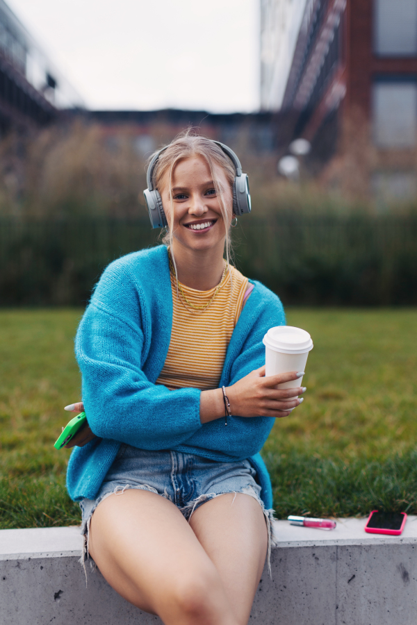 Portrait of a Generation Z girl student listening to music via wireless headphones outdoors in the city. Student spending free time alone, enjoying coffee in a reusable travel mug. Concept of gen Z as loneliest generation.