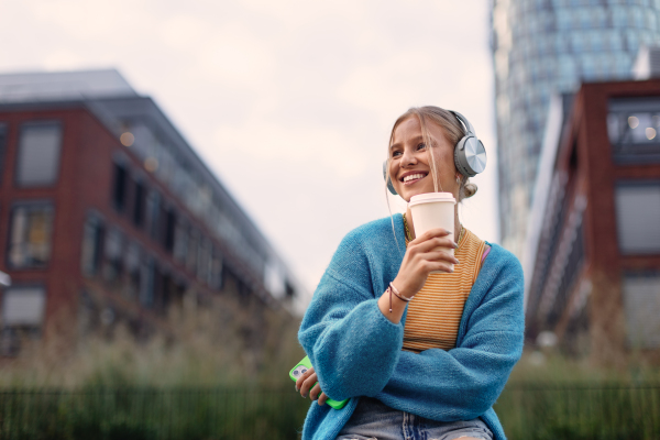 Portrait of a Generation Z girl student listening to music via wireless headphones outdoors in the city. Student spending free time alone, enjoying coffee in a reusable travel mug. Concept of gen Z as loneliest generation.