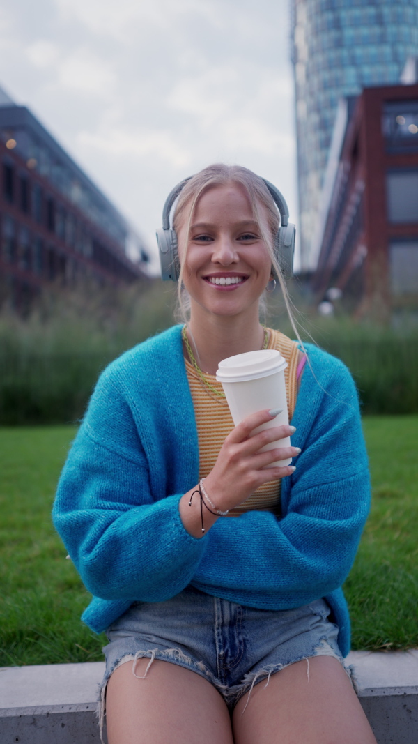 Video of a Generation Z girl student listening to music via wireless headphones outdoors in the city. Student spending free time alone, enjoying coffee in a reusable travel mug. Concept of gen Z as loneliest generation.