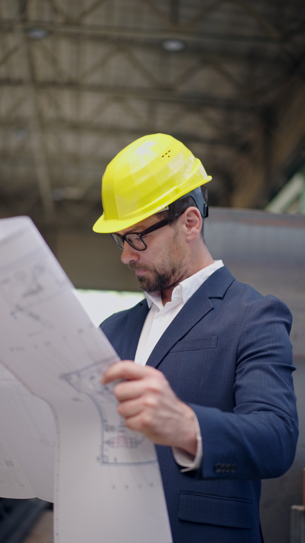 Vertical footage of engineer businessman with protective helmet in a factory, checking blueprints.