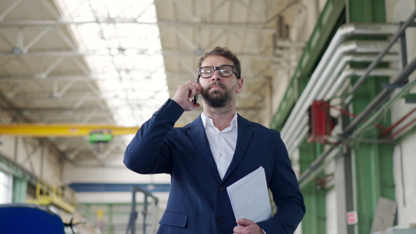 Engineer businessman with protective helmet in factory, holding blueprint and calling to somebody.