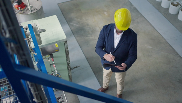 Engineer businessman with protective helmet in a factory, holding digital tablet and doing audit, high angle view.