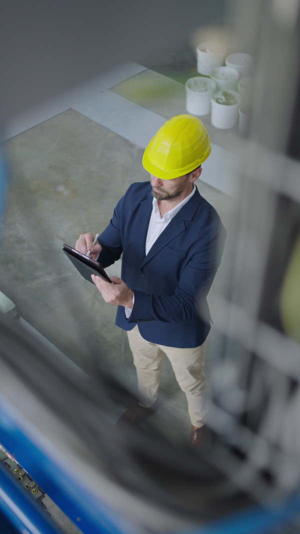 Engineer businessman with protective helmet in a factory, holding digital tablet and doing audit, vertical footage, high angle view.