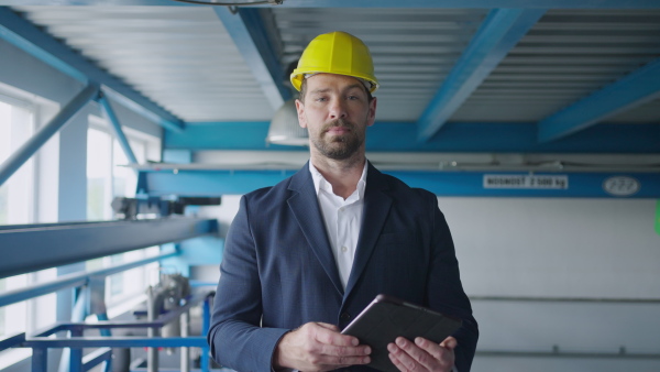 Engineer businessman with protective helmet in a factory, holding digital tablet and looking at camera.