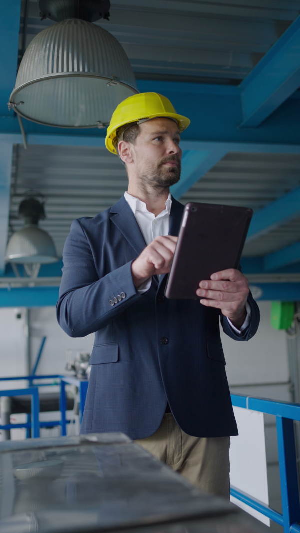 Vertical footage of an inpector in factory with protective helmet checking processes, doing audit with digital tablet.