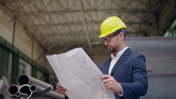 Engineer businessman with protective helmet in a factory, checking blueprints.