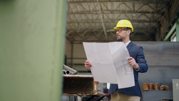 Engineer businessman with protective helmet in a factory, checking blueprints.