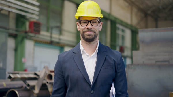 Engineer businessman with protective helmet in a factory, checking blueprints and looking at camera.