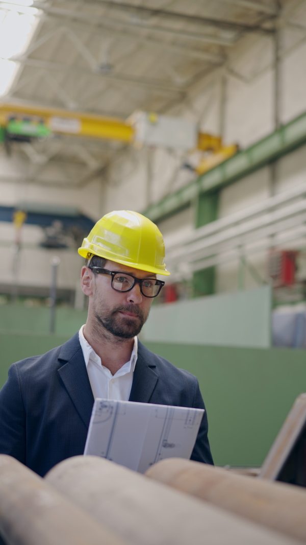 Vertical footage of engineer businessman with protective helmet in a factory, checking blueprints.