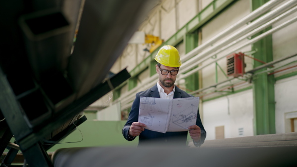 Engineer businessman with protective helmet in a factory, checking blueprints.