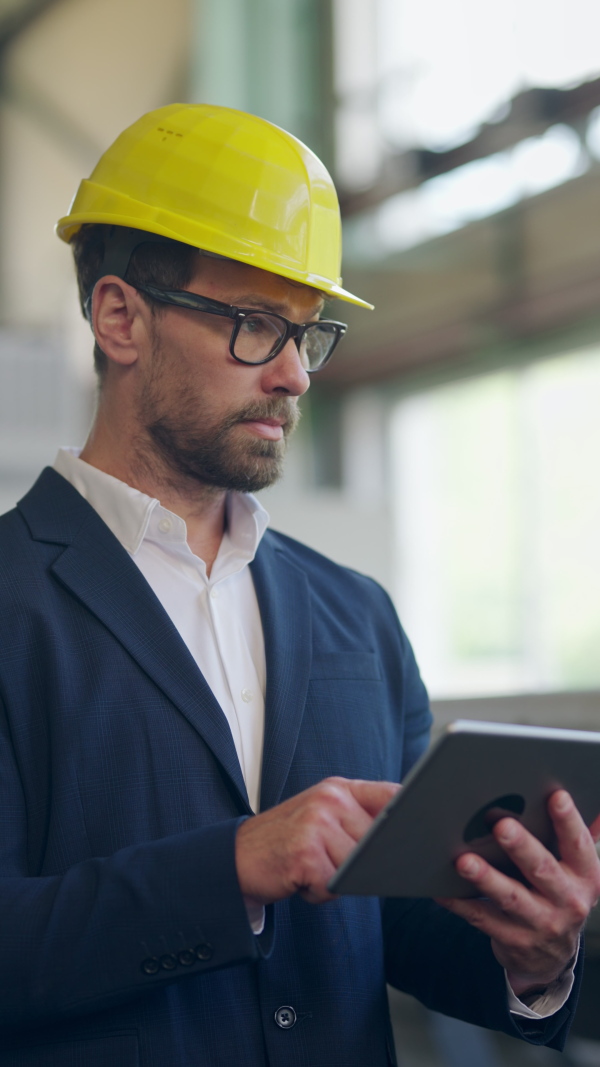 Engineer businessman with protective helmet in a factory, holding digital tablet and doing audit, vertical footage.