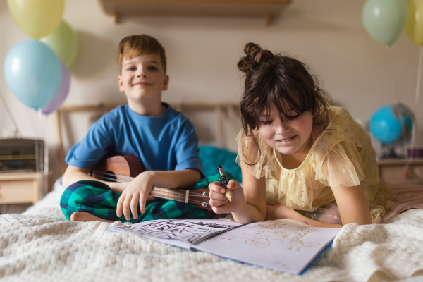 Little boy plaing on the guitar, while his sister drawing a picture on a bed.