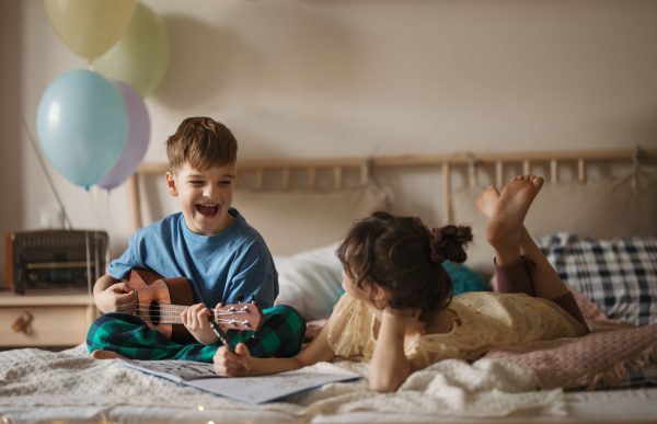 Little boy plaing on the guitar, while his sister drawing a picture on a bed.