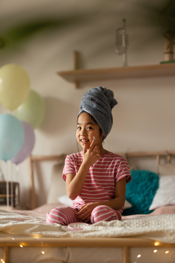 Little girl sitting on her bed and appling cream on her face.