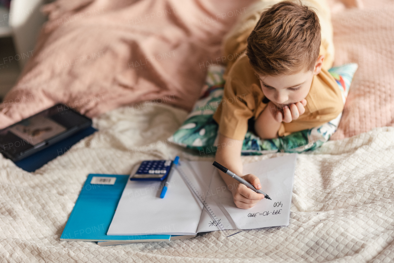 Little boy lying on a bed and doing the homework.