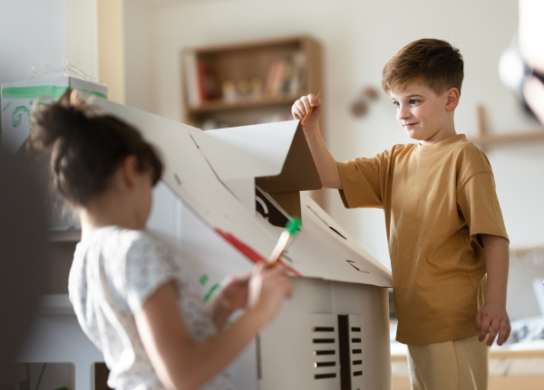 Happy children painting handmade carboard house together, working on school project. Creative indoor activities for kids.