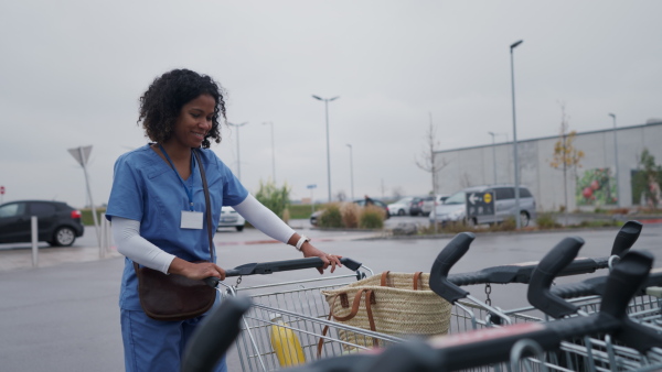 Female doctor grocery shopping after work. Beautiful healthcare worker pushing shopping cart with groceries across store parking lot. Work-life balance of healthcare worker as parent and partner.