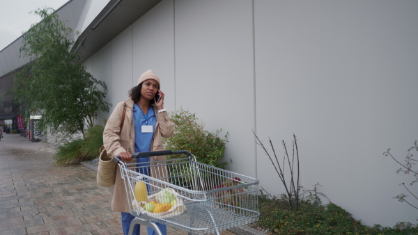 Female doctor grocery shopping after work. Beautiful healthcare worker pushing shopping cart with groceries across store parking lot. Work-life balance of healthcare worker as parent and partner.