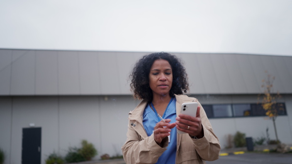 Female nurse having break at work, smoking cigarette and scrolling on smartphone. Healthcare worker taking break from work, resting outdoors, feeling exhausted.