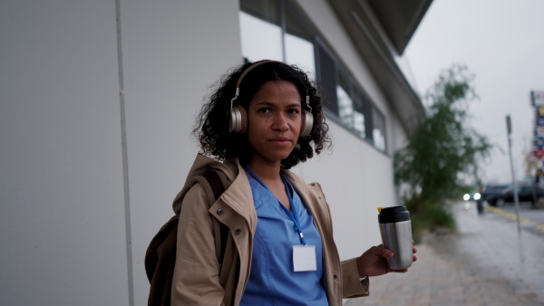 Female nurse, doctor going home from work, waiting for bus, listening music, drinking coffee and scrolling on smartphone. Work-life balance of healthcare worker as parent and partner.