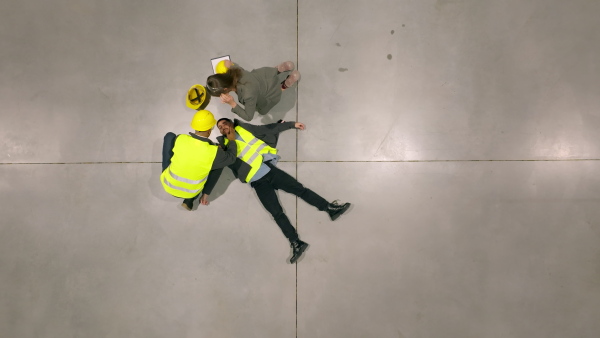 Top view of colleagues giving first aid to unconscious worker after accident at work in warehouse, modern factory. Calling the ambulance. Concept of occupational safety and health in workplace.