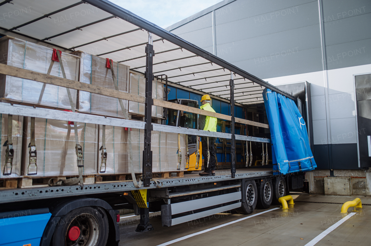 Side view of warehouse receiver standing inside of truck in cargo area, open trailer. Receiving clerk holding tablet, looking at cargo details, checking delivered items, goods against order.