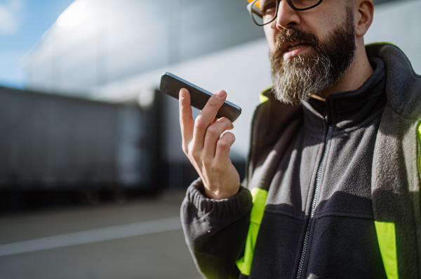 Side view of warehouse worker standing outdoors, phone calling with truck driver. Warehouse receiver waiting for a delivery.