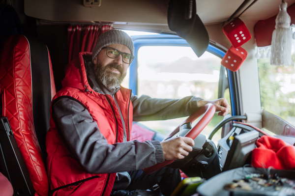 Portrait of confident professional truck driver sitting in a truck cabin, looking at camera and smiling. Inside of vehicle.