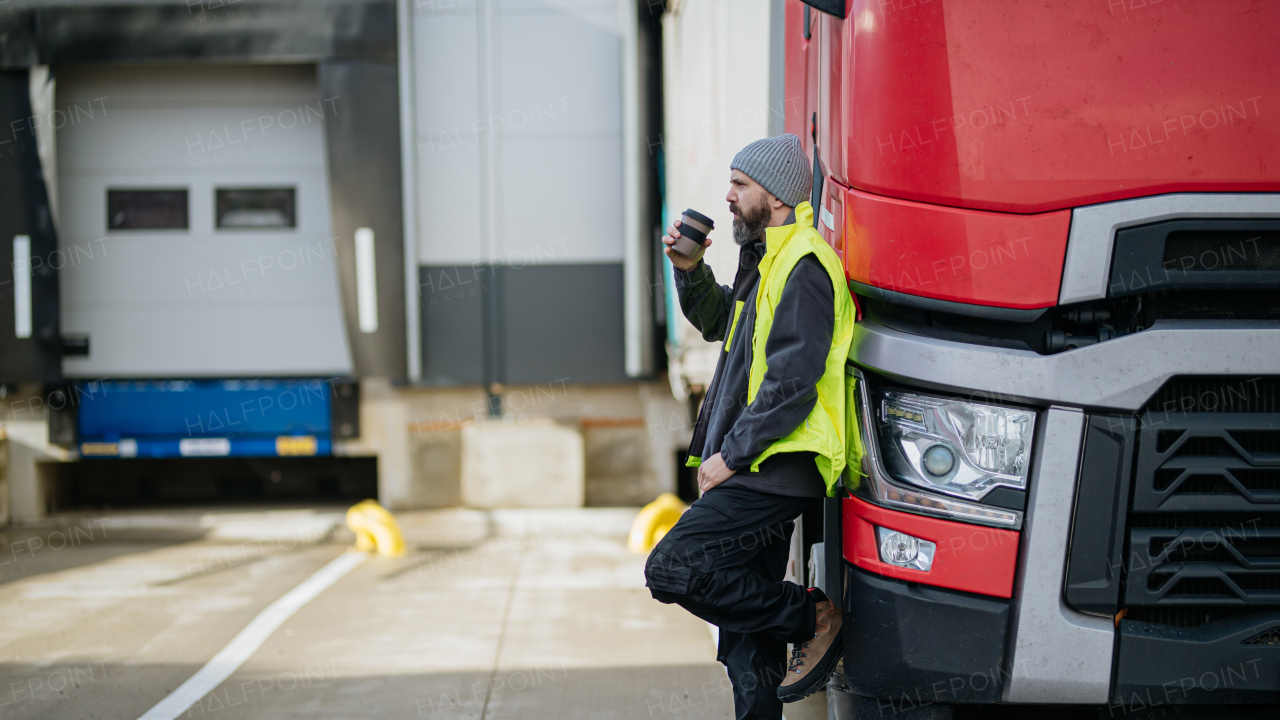 Truck driver leaning against hist red truck and drinking coffee, waiting for warehouse workers.