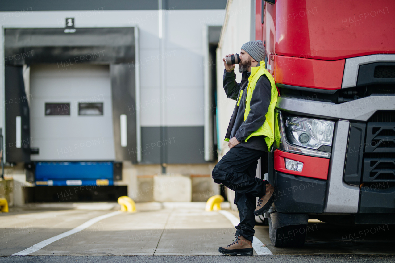 Truck driver leaning against hist red truck and drinking coffee, waiting for warehouse workers.