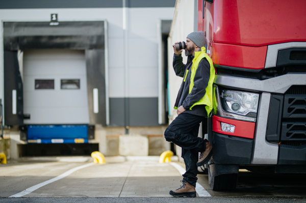 Truck driver leaning against hist red truck and drinking coffee, waiting for warehouse workers.