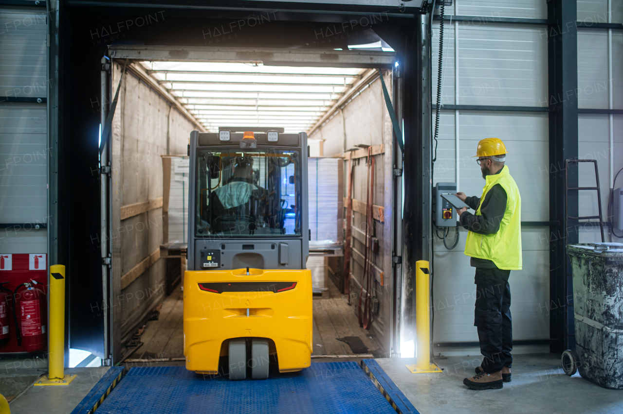 Warehouse receiver, clark overseeing the storing of delivered items, holding tablet, looking at cargo details. Forklift carrying pallets with goods.