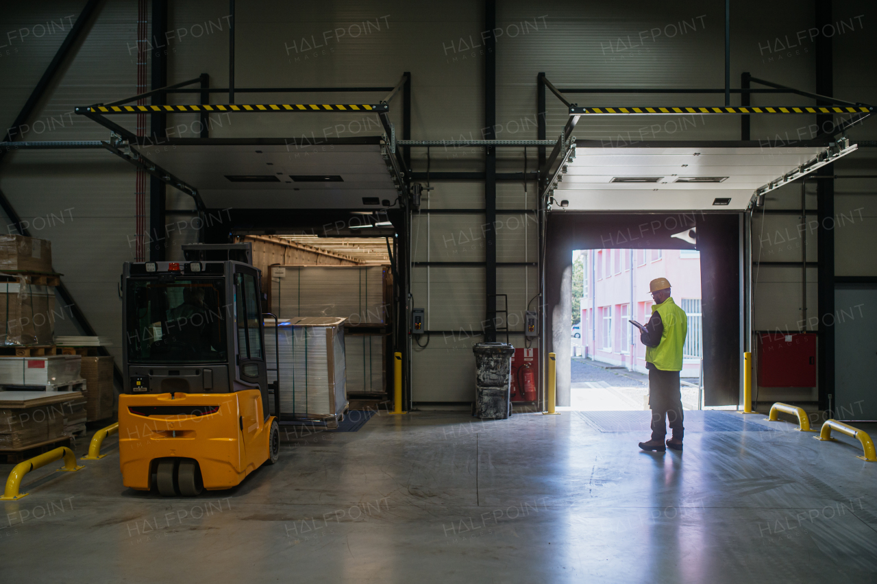 Warehouse receiver, clark overseeing the storing of delivered items, holding tablet, looking at cargo details. Forklift carrying pallets with goods.