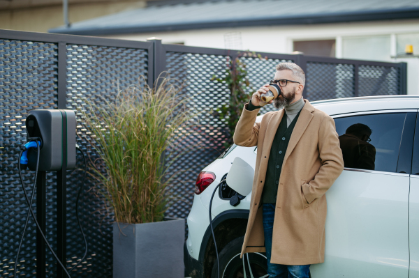 Portrait of businessman waiting while electric car is charging, leaning against the vehicle, scrolling on his smartphone, and drinking coffee. An electric vehicle charging station at home.