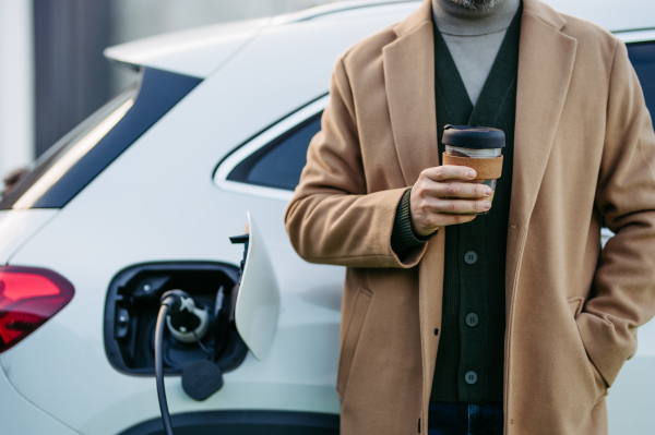 Close up of businessman in coat waiting while electric car is charging, leaning against the vehicle, drinking coffee. An electric vehicle charging station at home.