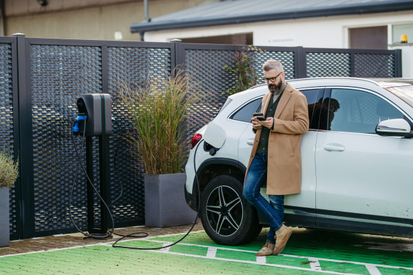 Portrait of businessman waiting while electric car is charging, leaning against the vehicle, scrolling on his smartphone, and drinking coffee. An electric vehicle charging station at home.