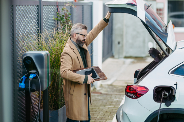 Businessman charging electric car before going to office. An electric vehicle charging station in front of the office building. Charging at work, workplace.