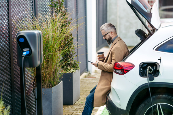 Portrait of businessman waiting while electric car is charging, sitting in car trunk, scrolling on his smartphone, and drinking coffee. An electric vehicle charging station at home.