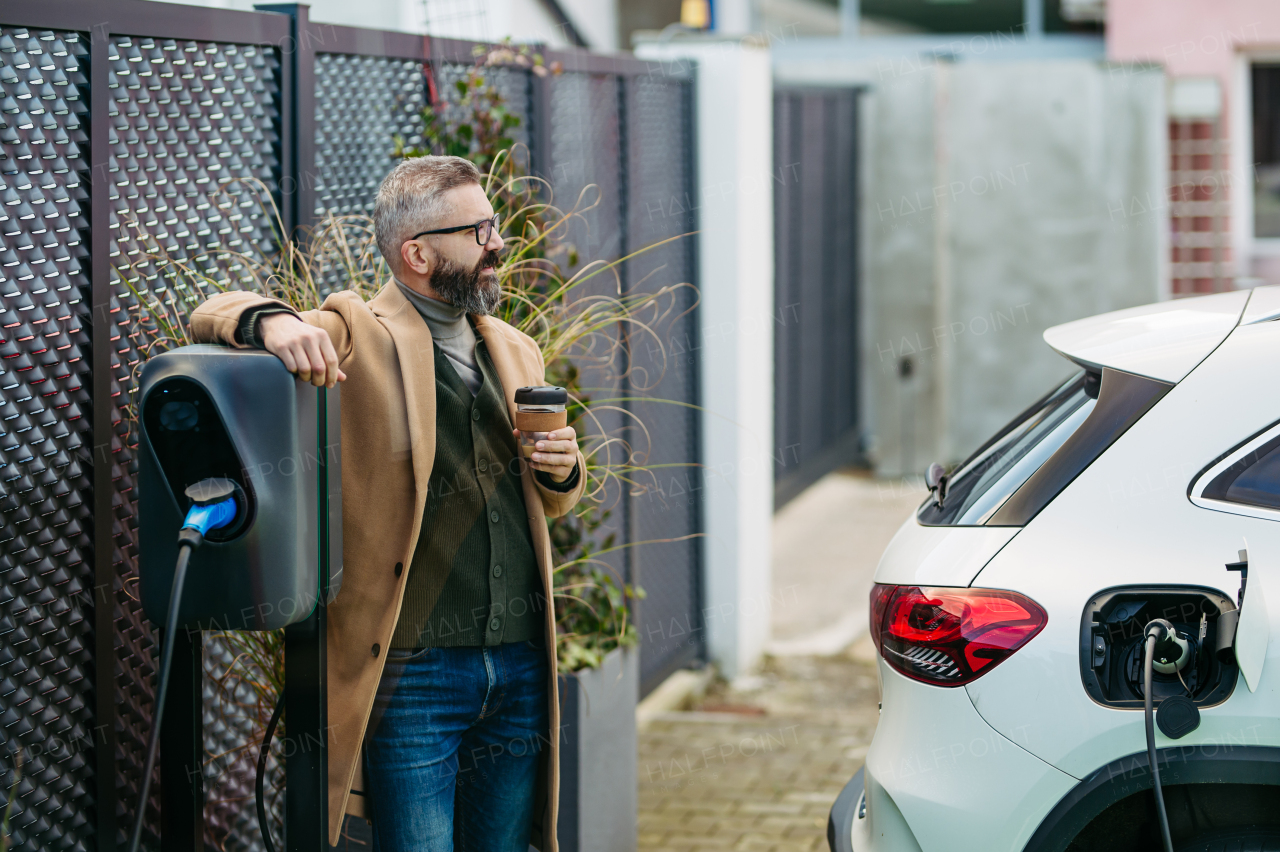 Portrait of businessman waiting while electric car is charging, leaning against the vehicle, scrolling on his smartphone, and drinking coffee. An electric vehicle charging station at home.