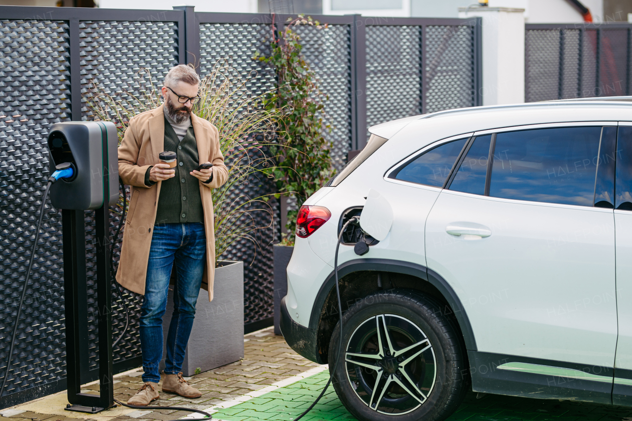 Portrait of businessman waiting while electric car is charging, leaning against the vehicle, scrolling on his smartphone, and drinking coffee. An electric vehicle charging station at home.