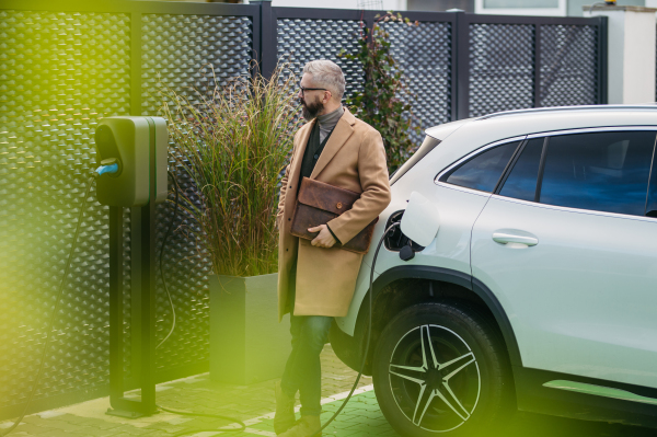 Businessman charging electric car before going to office. An electric vehicle charging station in front of the office building. Charging at work, workplace.