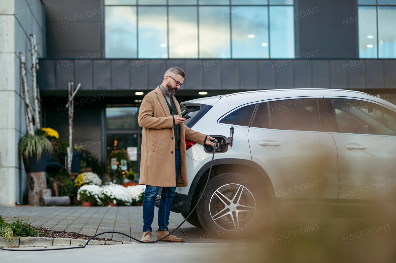 Man plugging in charging plug in electric car before going to shopping. An electric vehicle charging station in front of shop building, supermarket. Charging while shopping.