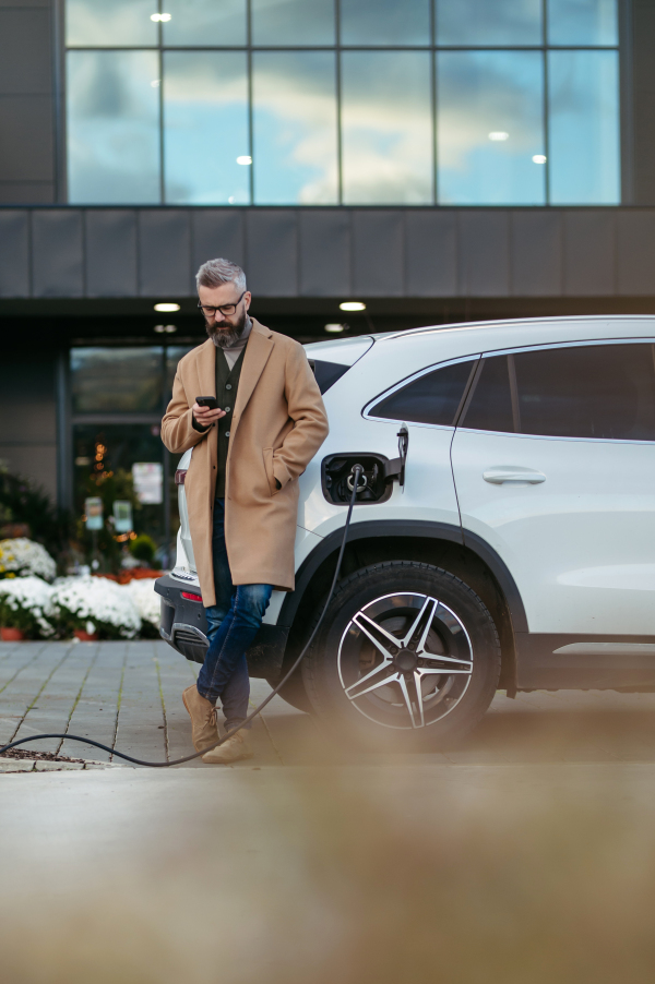 Man charging his electric car before going to shopping. A public charging station in front of shop building, supermarket. Charging while shopping.