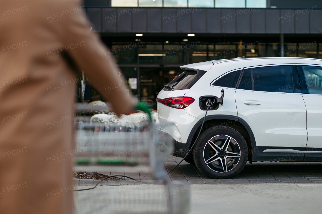 Man charging his electric car before going to shopping. A public charging station in front of shop building, supermarket. Charging while shopping.