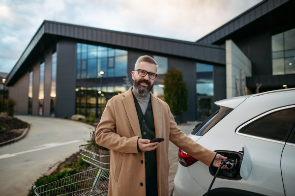 Man plugging in charging plug in electric car before going to shopping. An electric vehicle charging station in front of shop building, supermarket. Charging while shopping.